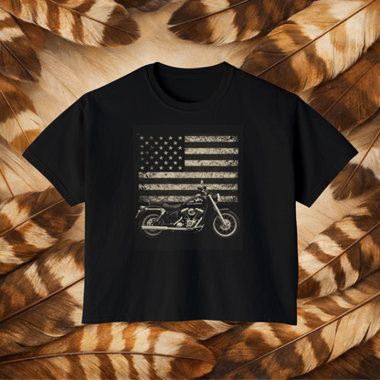 A black short-sleeve crewneck t-shirt featuring a distressed white graphic of an American flag and a motorcycle, displayed flat against a background of brown and tan feathers.