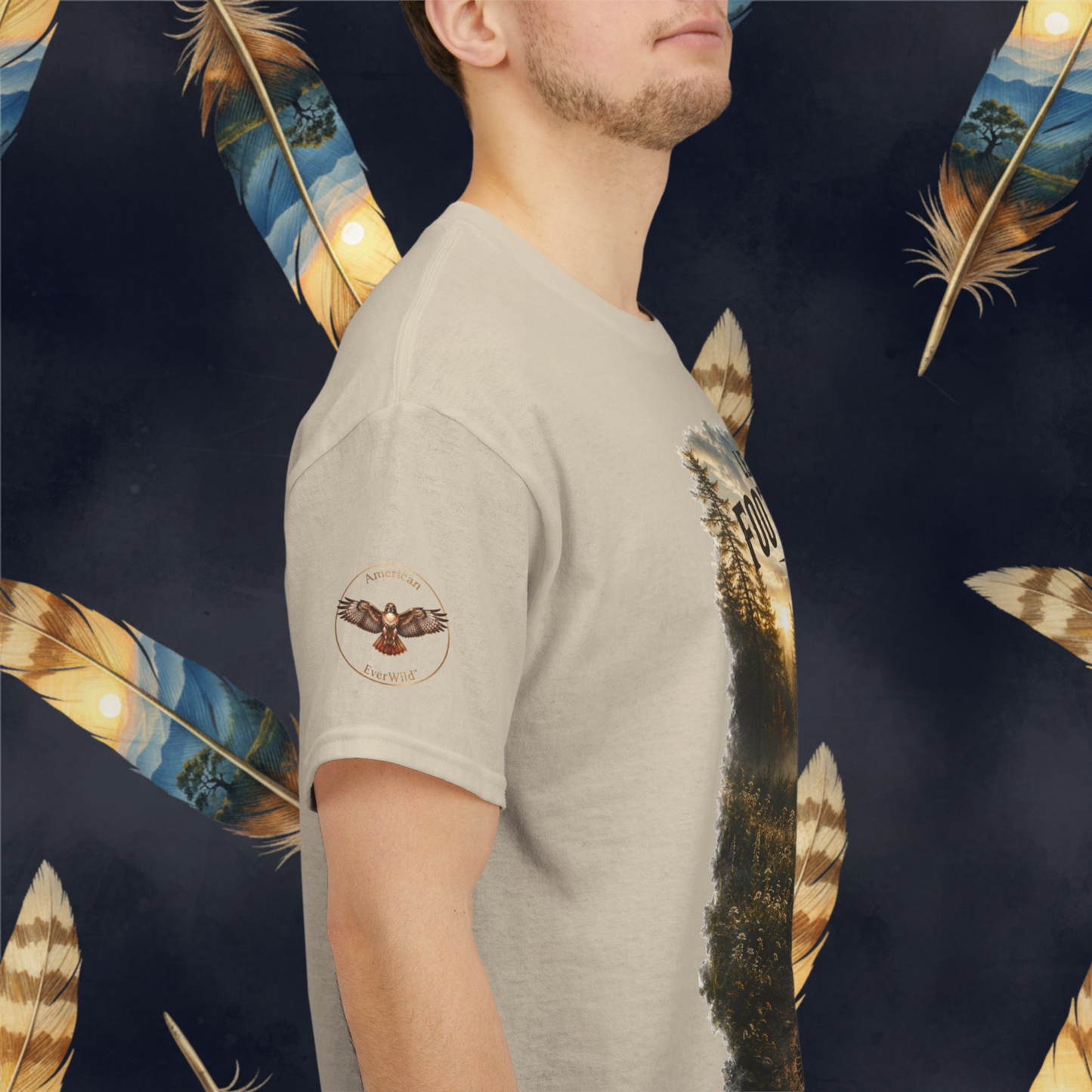 A man wearing a sand color crewneck t-shirt with a graphic print of an hawk and 
"American EverWild" inside a golden brown circle,  shown from the side against a dark background with large decorative feathers.