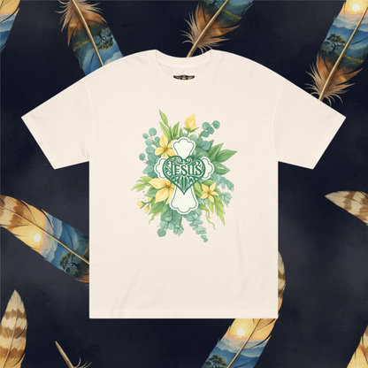 An cream color crewneck t-shirt featuring a central graphic of a green shamrock cross with the word 'JESUS' in stylized lettering, surrounded by watercolor green leaves, yellow flowers, and blue berries. The t-shirt is displayed flat against a dark background decorated with large golden-brown feathers.