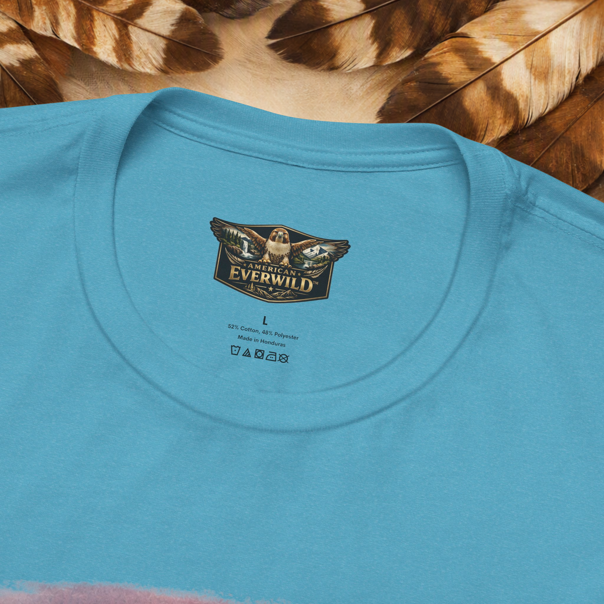 A close-up of a light blue crewneck t-shirt laid flat on a background of brown and beige feathers. The inside of the collar features a printed graphic logo for 'American EverWild' with a red-tailed hawk with painted wings.