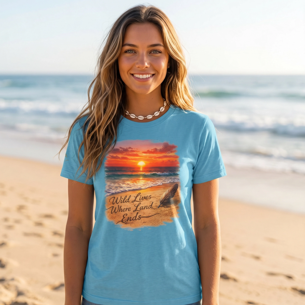 A woman standing on a sandy beach wearing a light blue graphic T-shirt with a sunset print and the text 'Wild Lives When Land Ends.'.