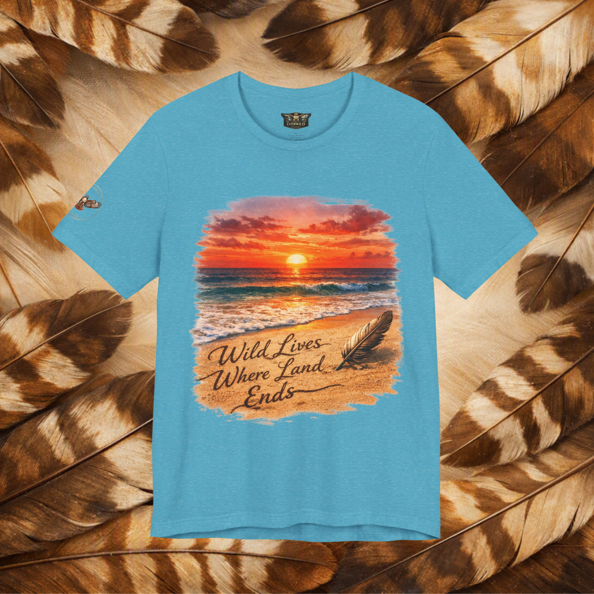 A teal tee shown from the front, featuring a ocean at sunset graphic with the text 'Wild Lives Where Land Ends'. The tee is displayed flat against a background of brown and beige feathers.