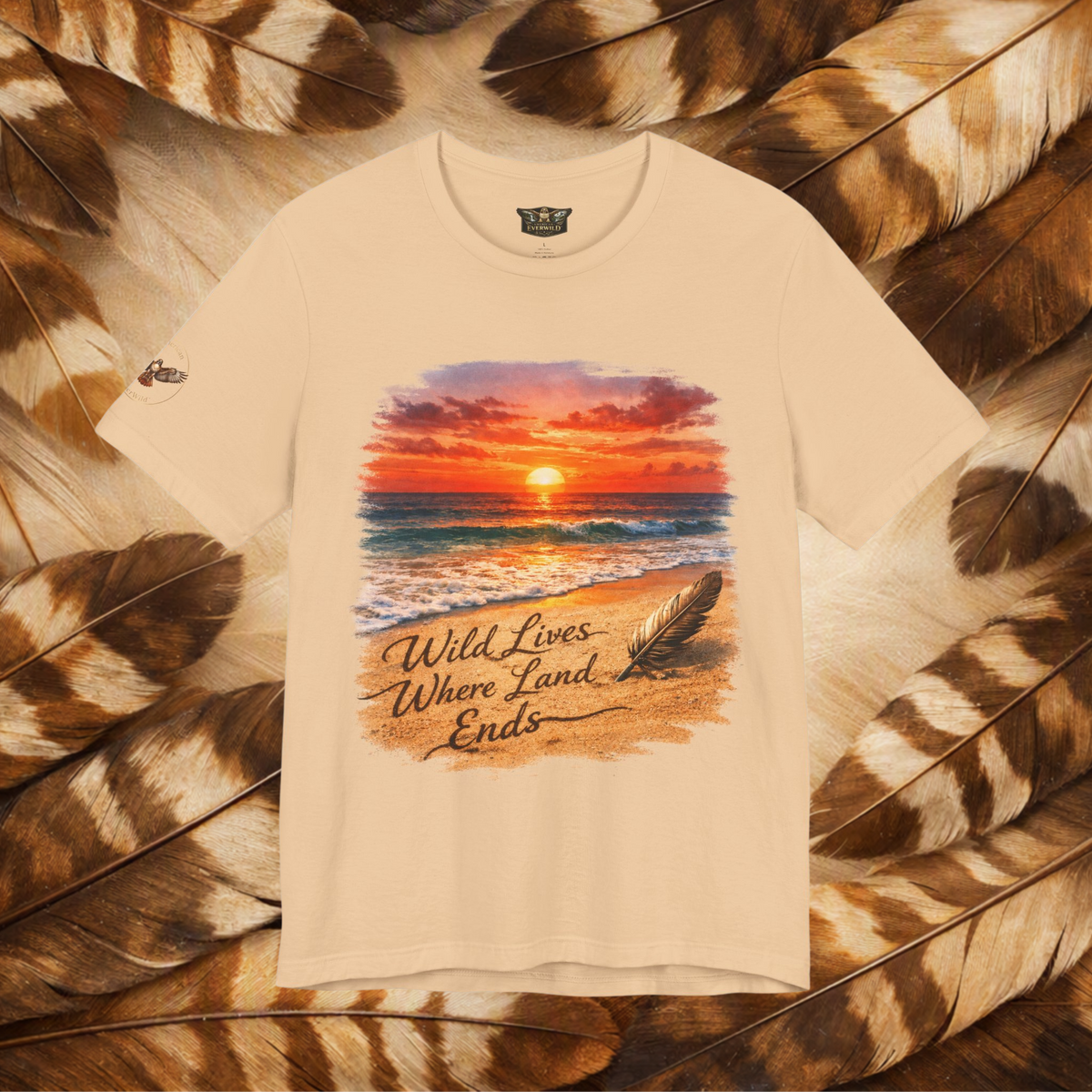 A cream-colored short-sleeve t-shirt with a colorful beach sunset graphic and the text 'Wild Lives Where Land Ends' printed on the front, displayed flat on a background of brown and beige feathers.