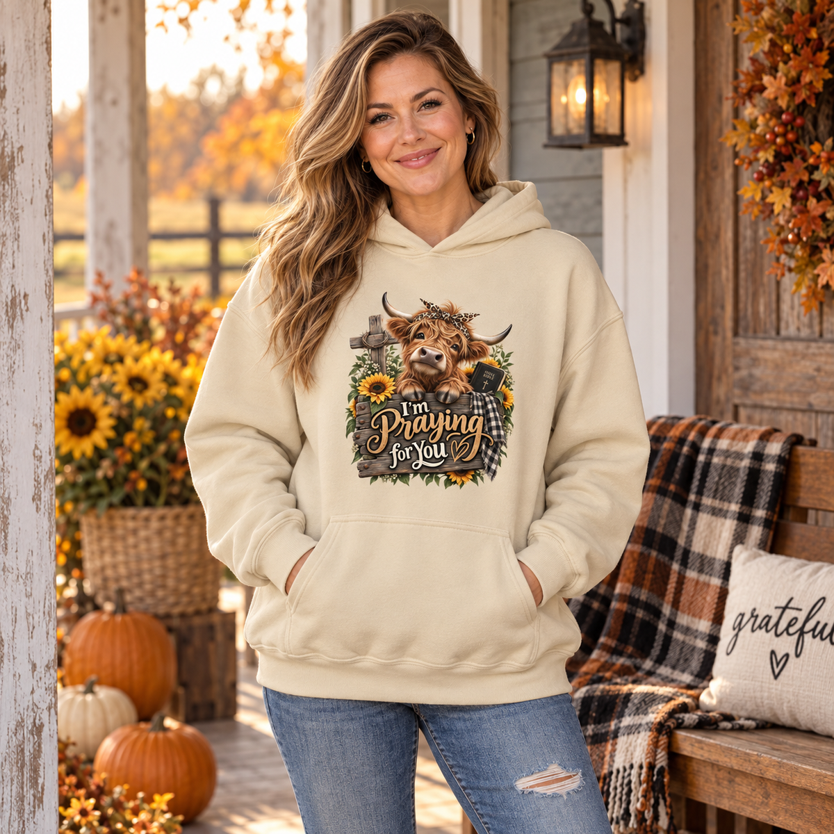 Woman on a front porch with her hands in the front pocket of a sand-colored hoodie with a playful country cow leaning over a fence with a bow in its hair, yellow flowers and a cross with the text, " I'm Praying for You" on the front chest.