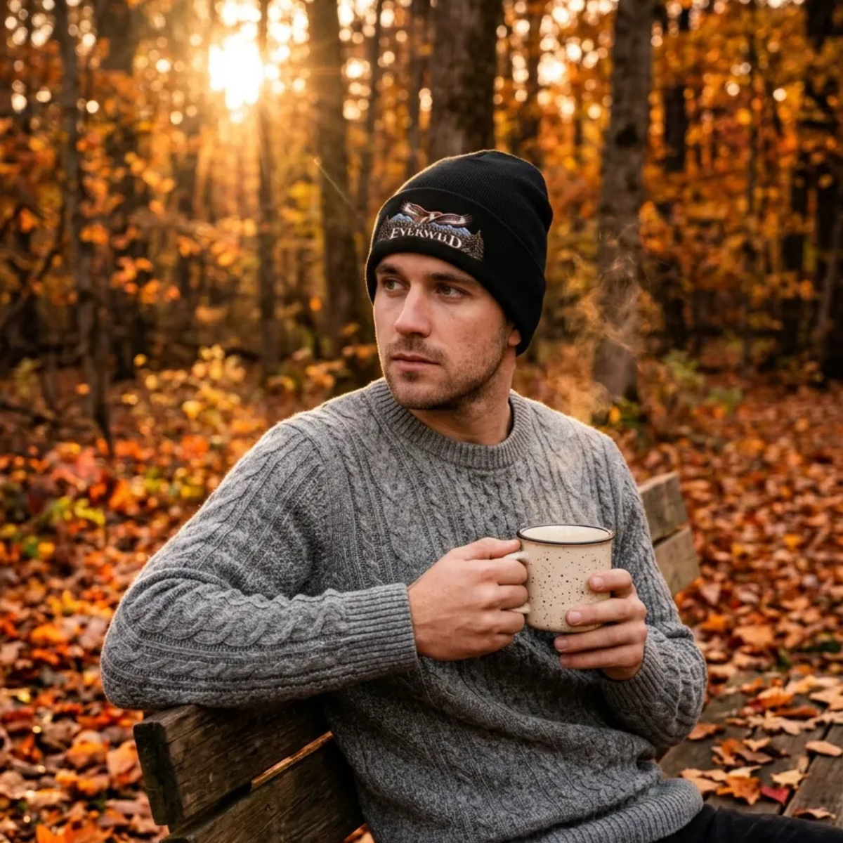 Man sitting in fall woods on a bench with a coffee mug. Man is wearing a Black American EverWild beanie and sweater.