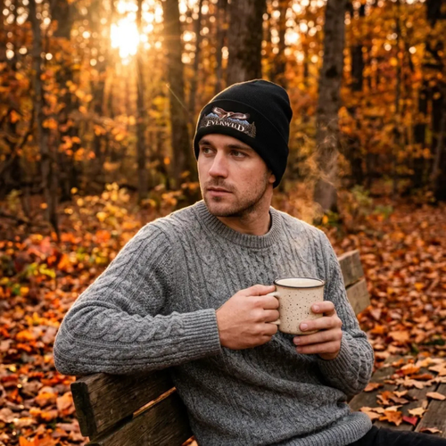 Man sitting in fall woods on a bench with a coffee mug. Man is wearing a Black American EverWild beanie and sweater.