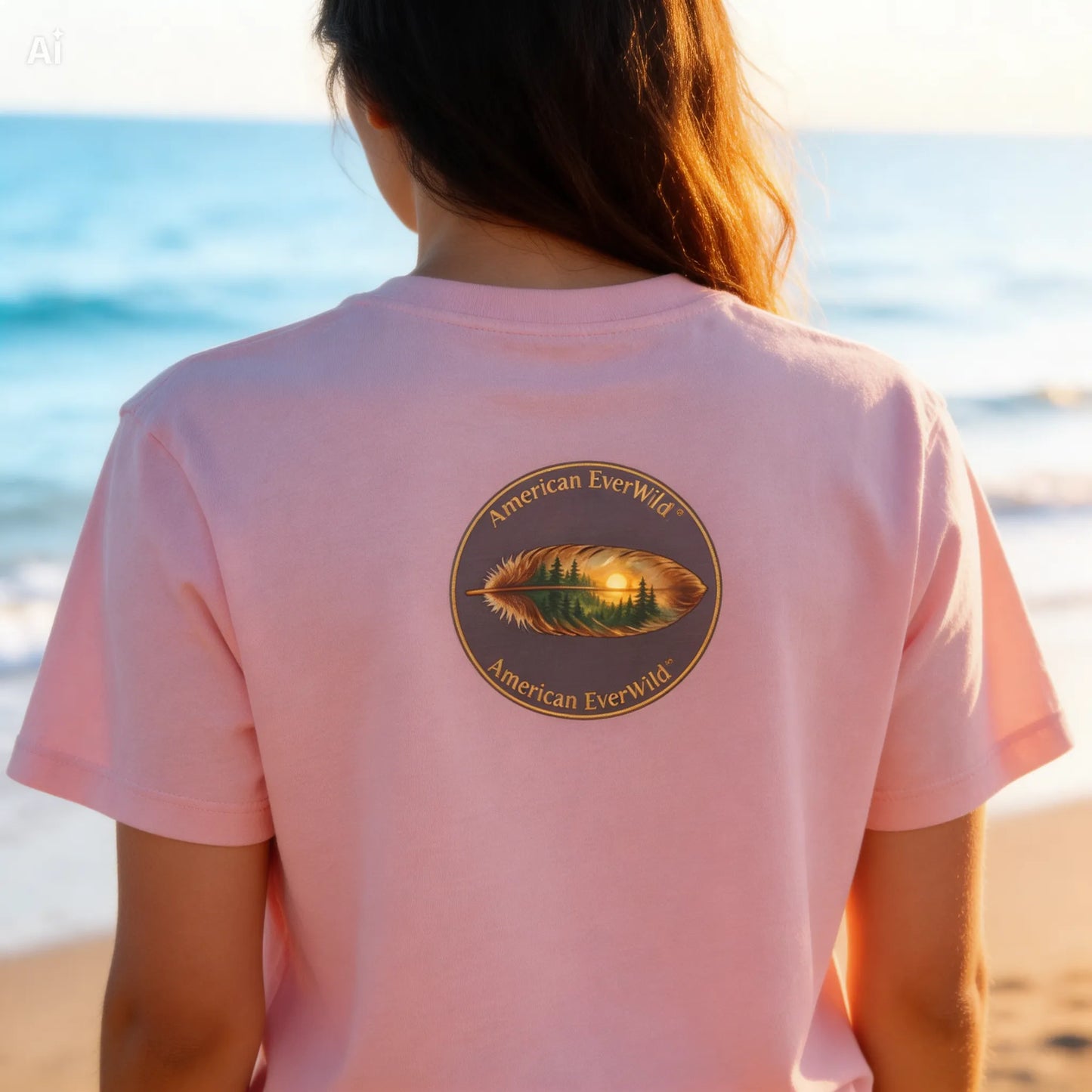 A woman viewed from behind wearing a light pink t-shirt with a circular American EverWild feather graphic on the back, set against a beach background at sunset.