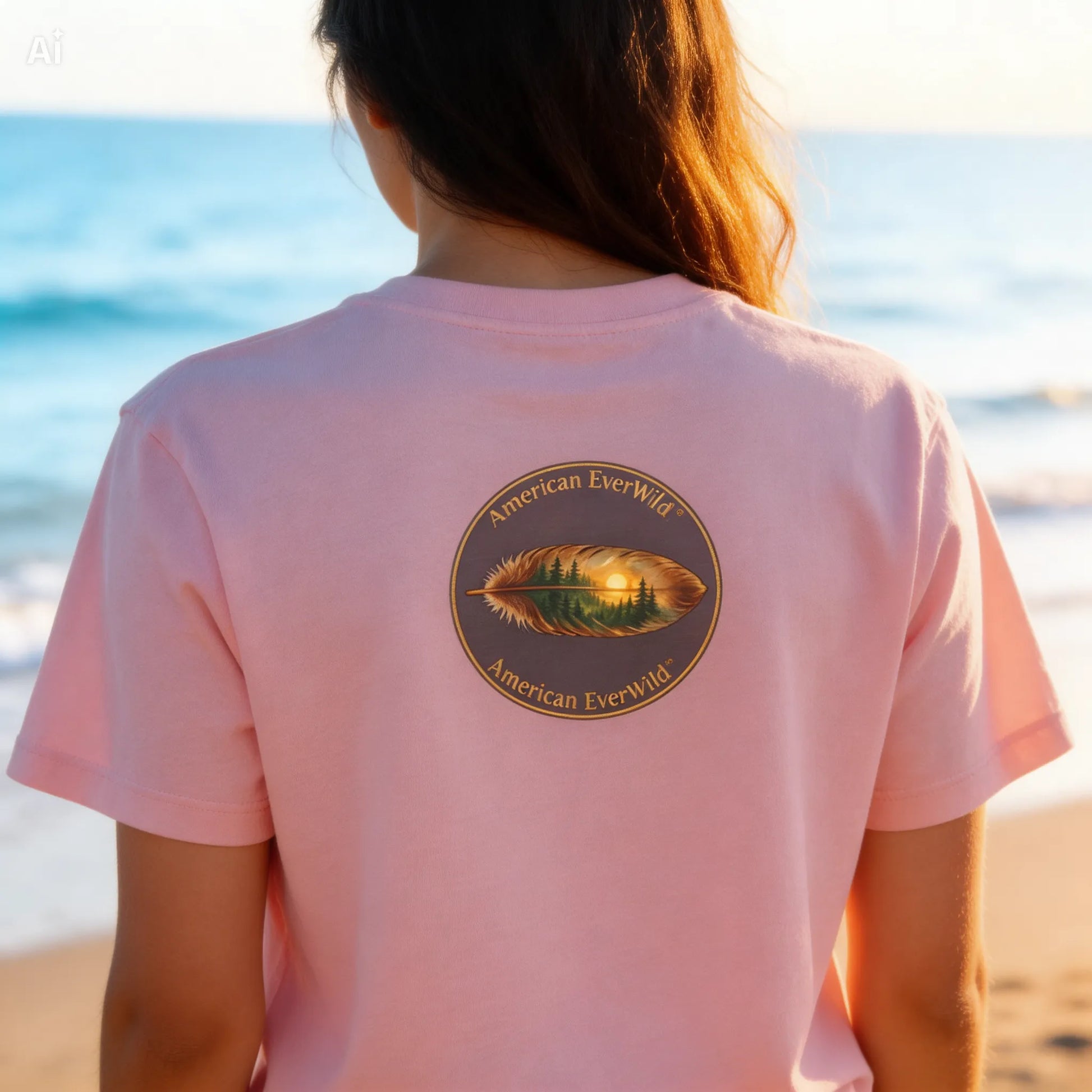 A woman viewed from behind wearing a light pink t-shirt with a circular American EverWild feather graphic on the back, set against a beach background at sunset.