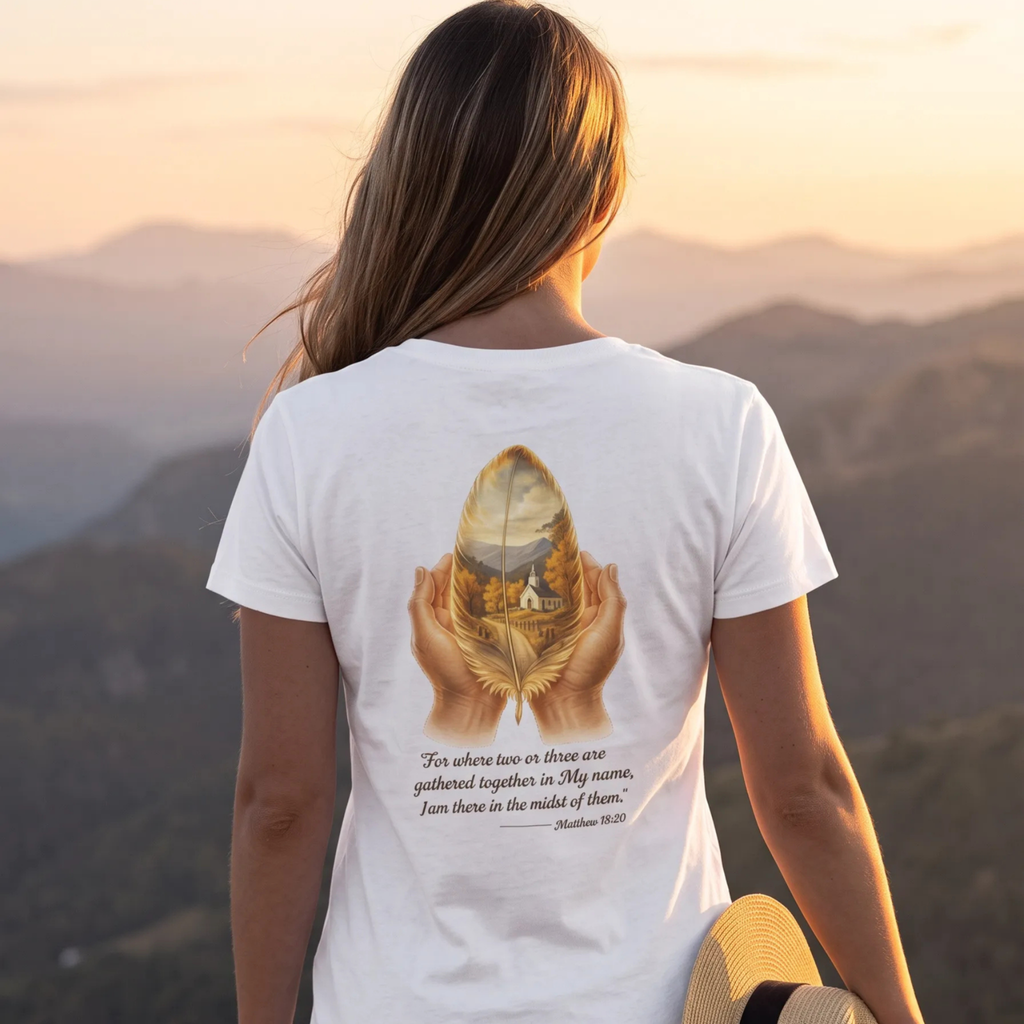 A woman viewed from behind wearing a white graphic t-shirt with a detailed print of hands holding a golden feather with a mountain landscape inside, and a Bible verse printed below. She is standing outdoors in front of a scenic mountain range at sunset.