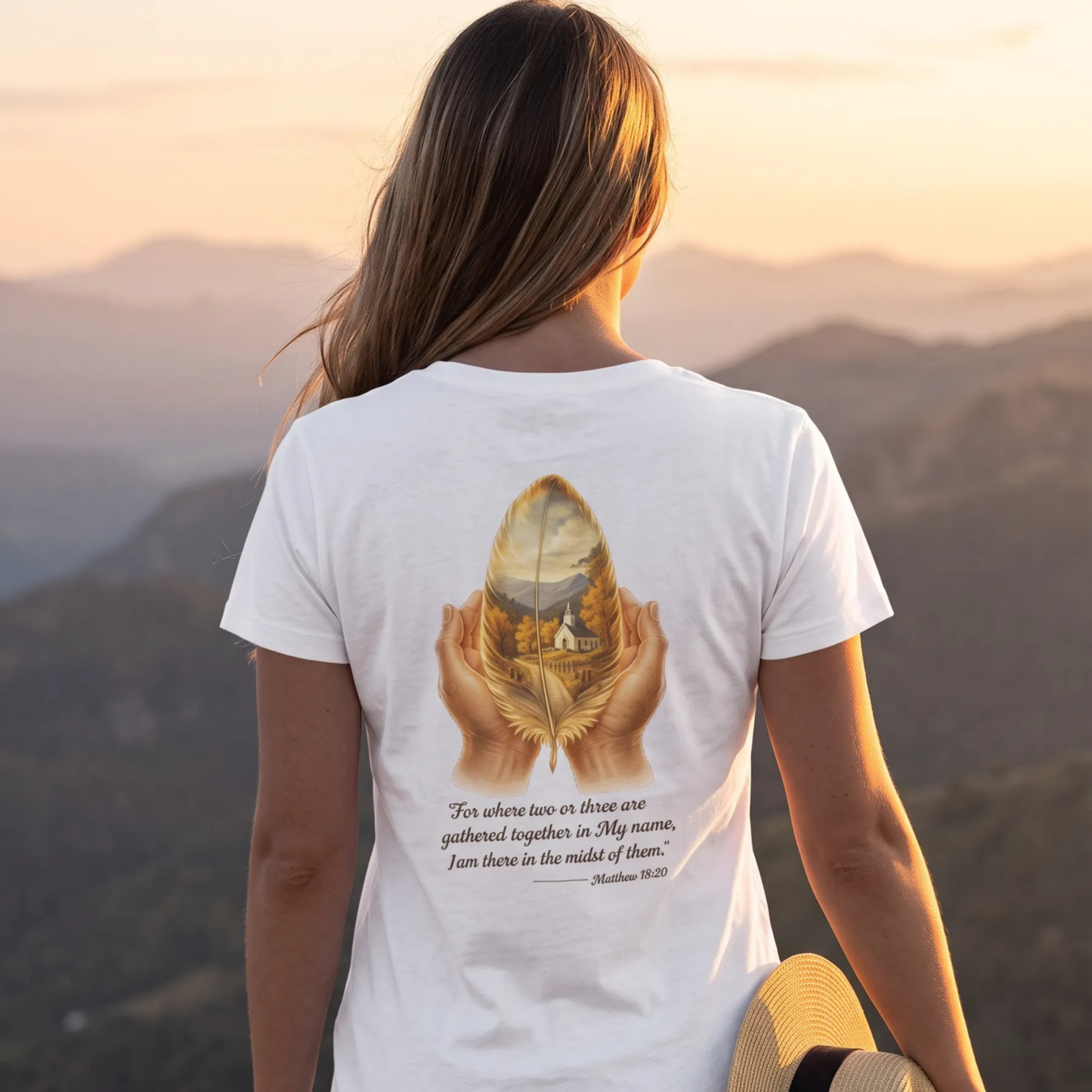 A woman viewed from behind wearing a white graphic t-shirt with a detailed print of hands holding a golden feather with a mountain landscape inside, and a Bible verse printed below. She is standing outdoors in front of a scenic mountain range at sunset.