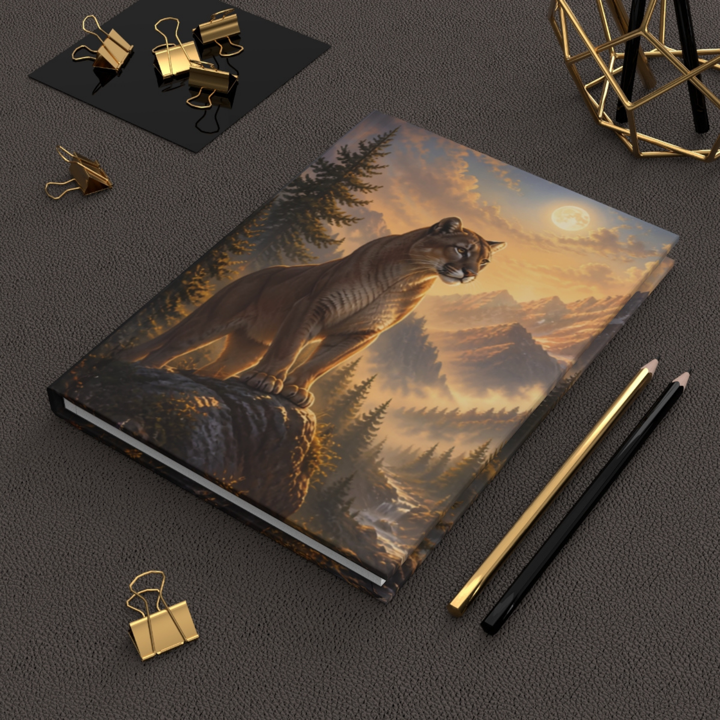 A hardcover notebook featuring a detailed illustration of a mountain lion in a pine forest at sunset, styled on a dark textured surface with gold and black office accessories.